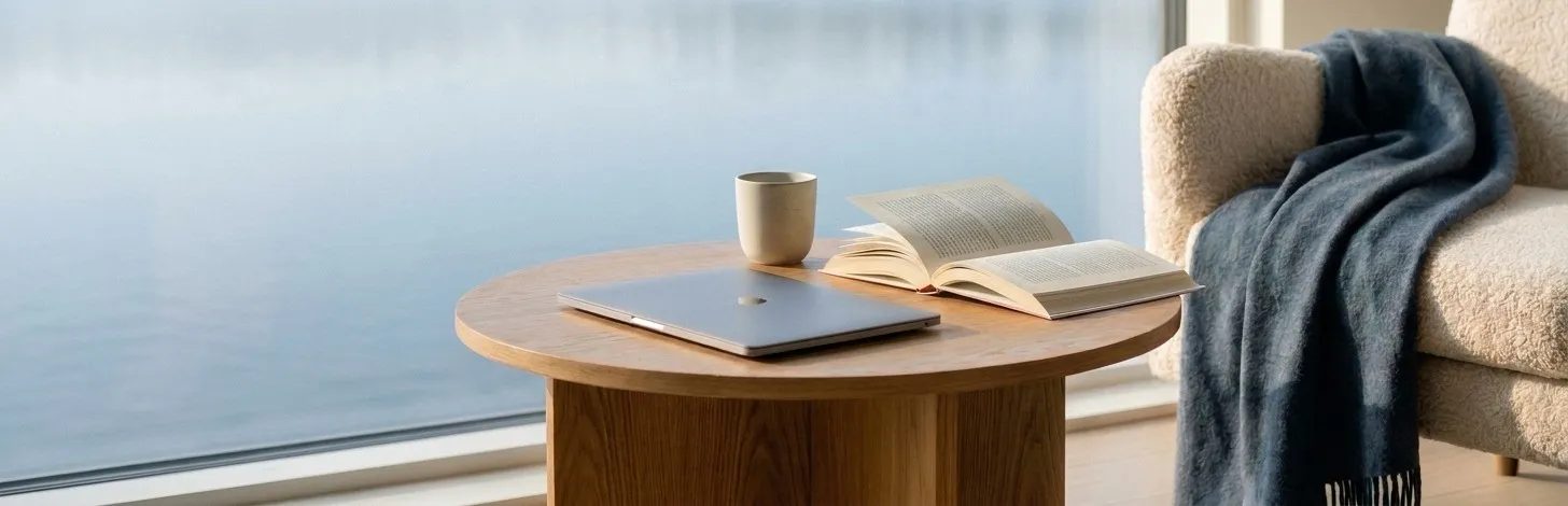 Zugeklappter Laptop, aufgeschlagenes Buch und Tasse auf einem runden Holztisch am Fenster mit Blick auf ruhiges Wasser